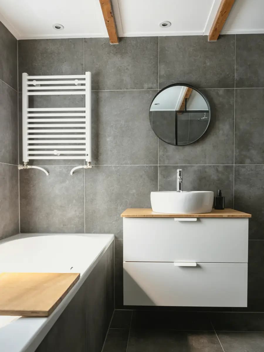 A tiled bathroom with large grey wall tiles, a white towel radiator, a wall-hung vanity unit with round mirror and a bath at the edge of the frame