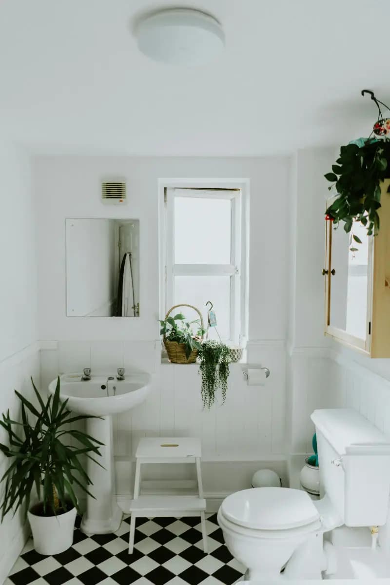 A white bathroom with a pedestal basin, a high-level cistern toilet, panelled walls, a wall extractor grille and a black and white chequered floor
