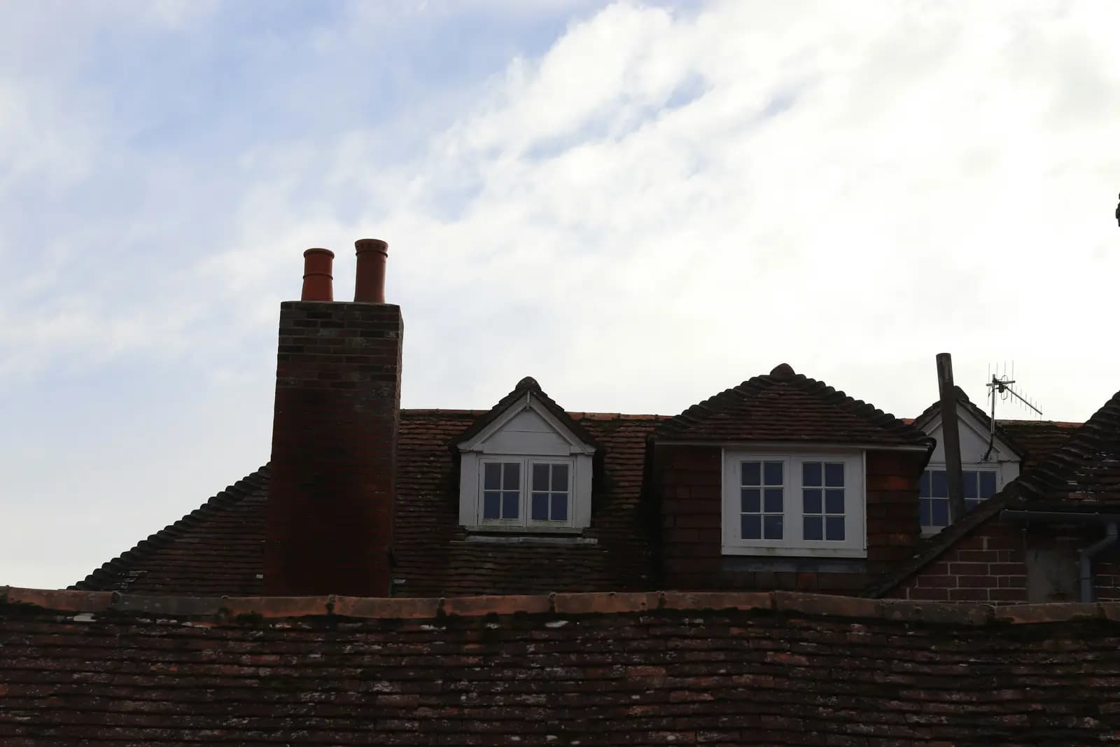 Weathered clay-tile roofs with a tall brick chimney stack and two white-framed dormer windows, one with a pitched roof and one with a hipped roof, against a cloudy sky