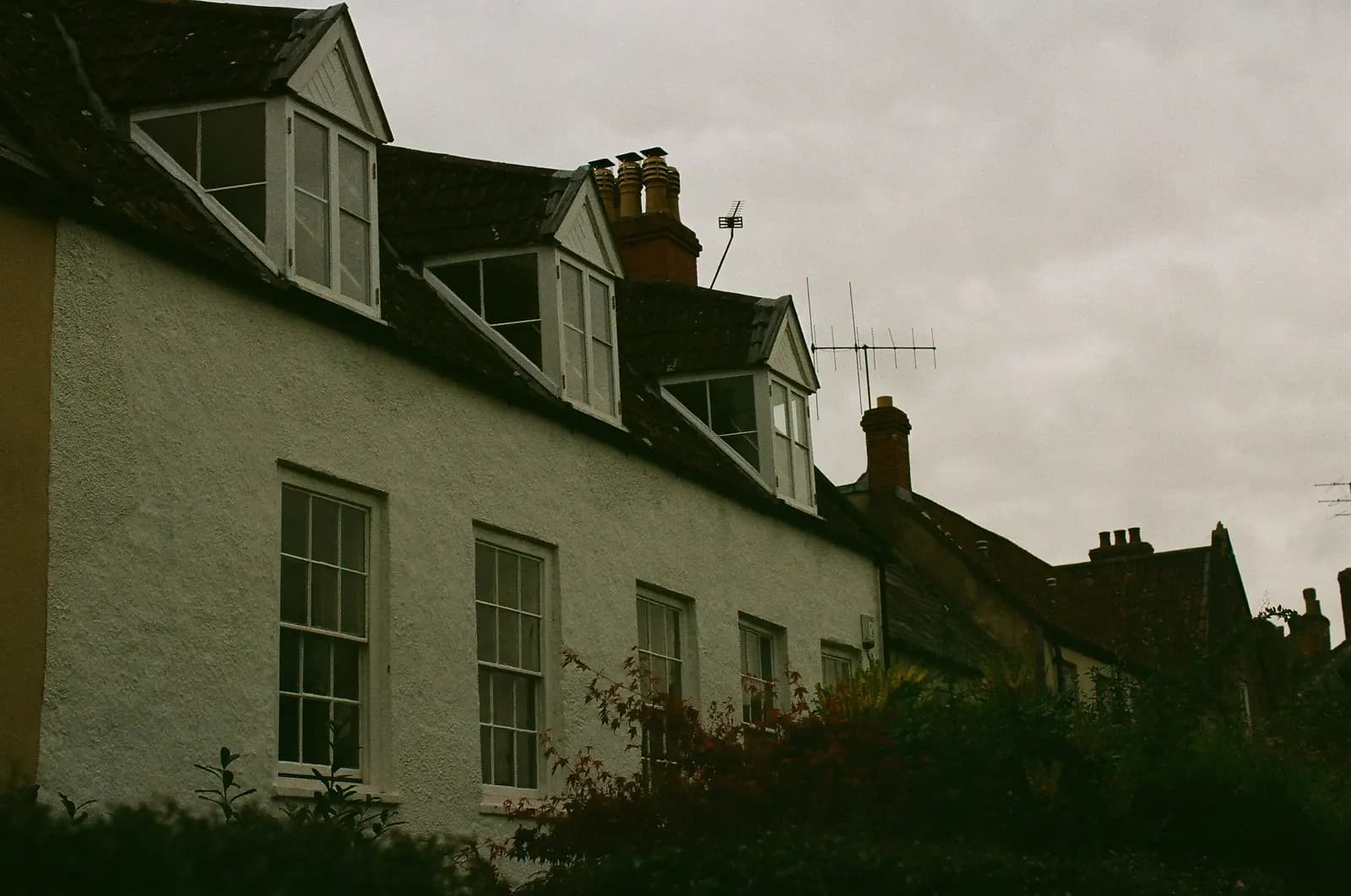 A white-rendered house with three gabled dormer windows set into a clay-tiled roof, chimney stacks and television aerials behind, under a grey English sky