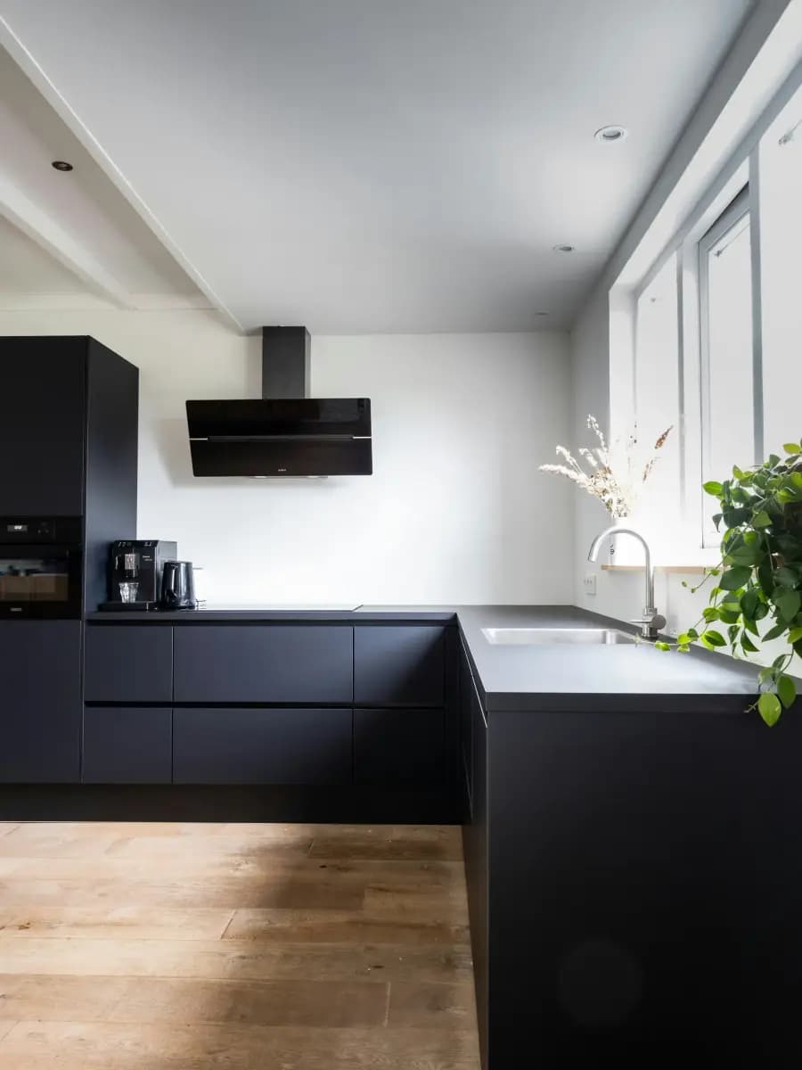 A modern kitchen with matte black base units, a light worktop, a tap under a window and a wooden floor