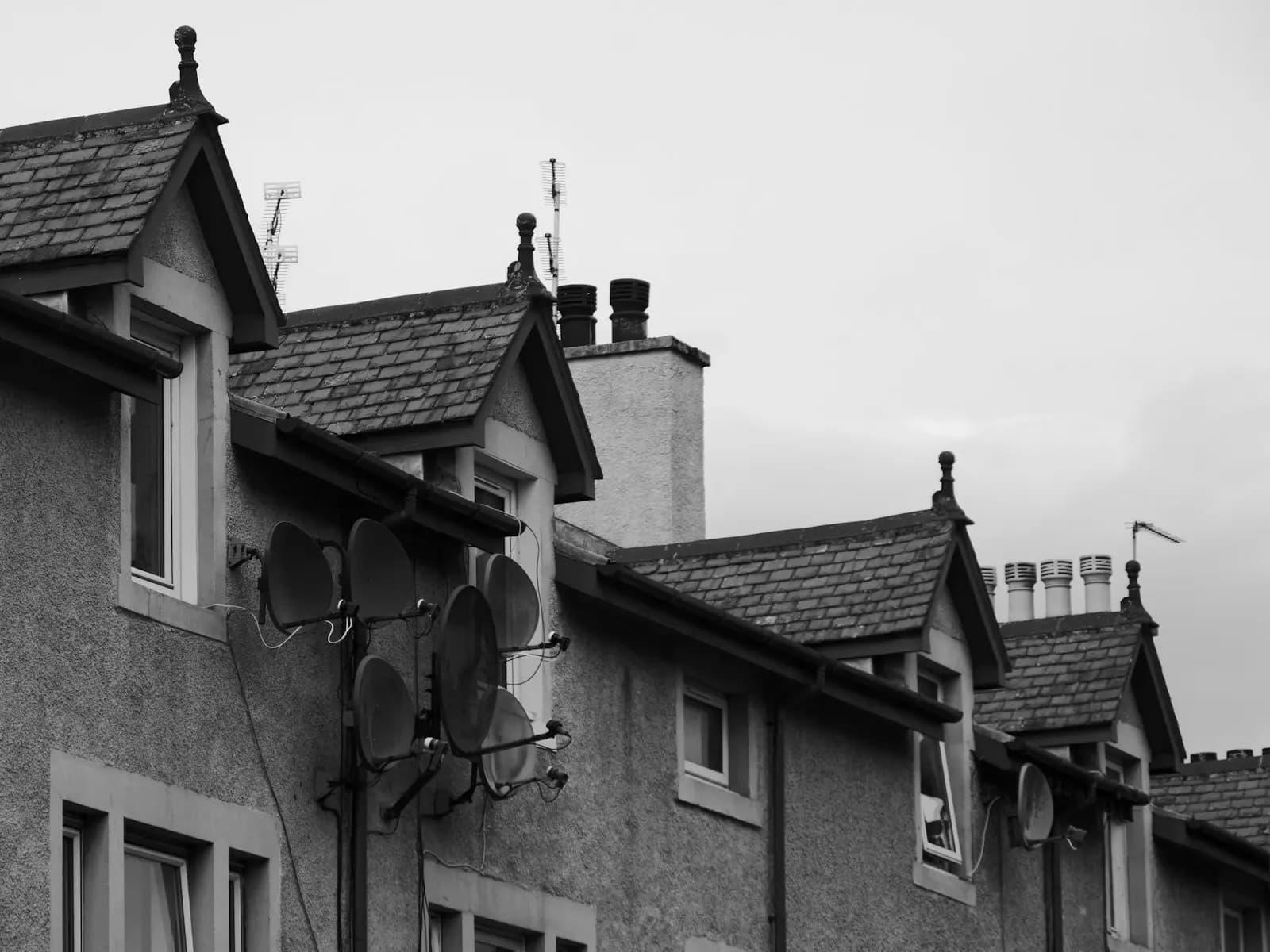 A black and white photo of a row of slate-roofed stone-and-render houses with pitched dormer windows, chimneys and satellite dishes, in Inverness