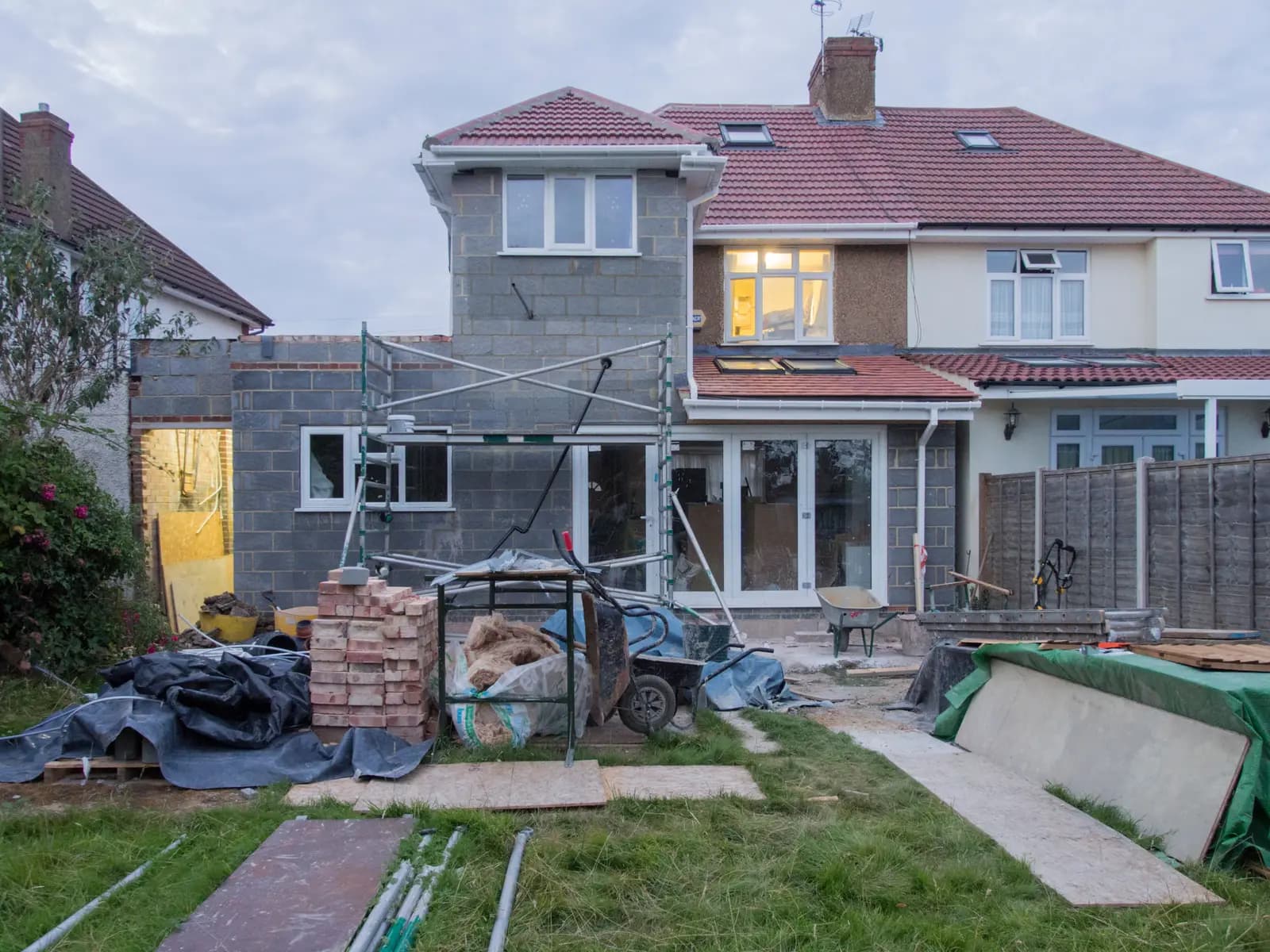 The rear of a semi-detached house with a part-built grey blockwork extension, scaffold tower, stacked bricks, wheelbarrows and a tiled roof with rooflights