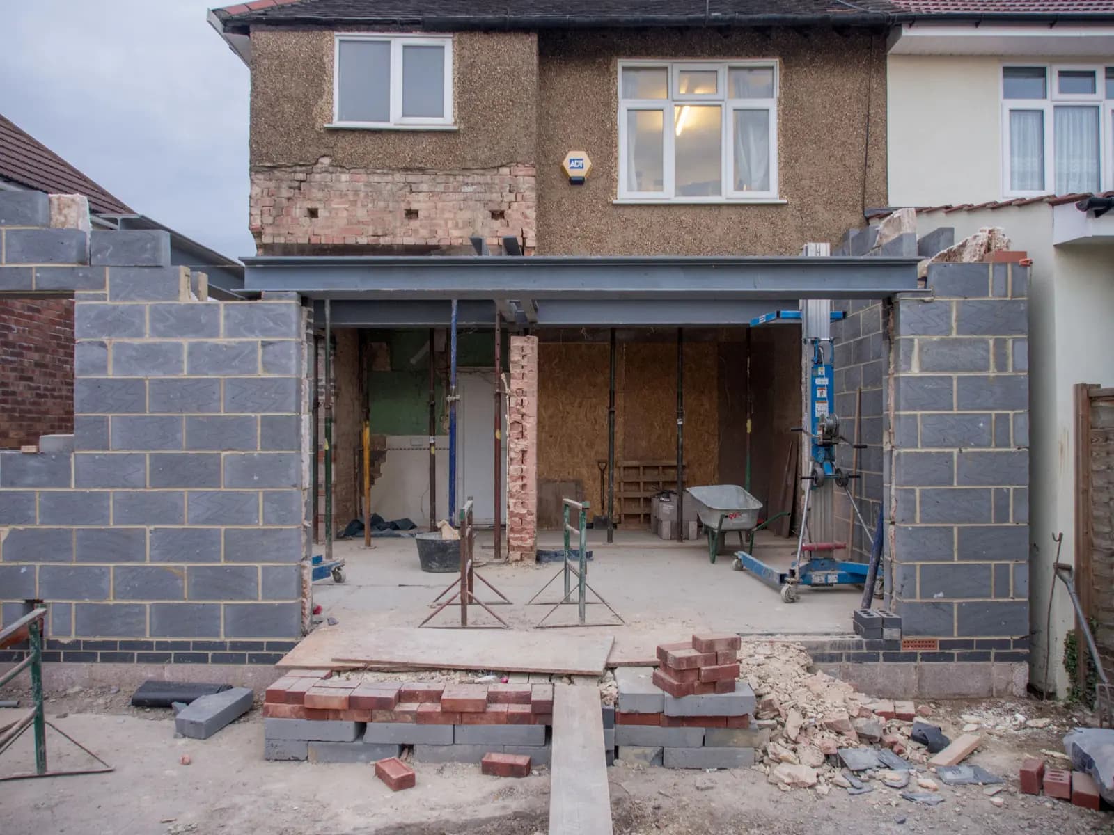 The back of a pebbledashed semi-detached house during an extension build, with a steel beam spanning the opened-up ground floor, adjustable props, a materials hoist and new blockwork walls either side