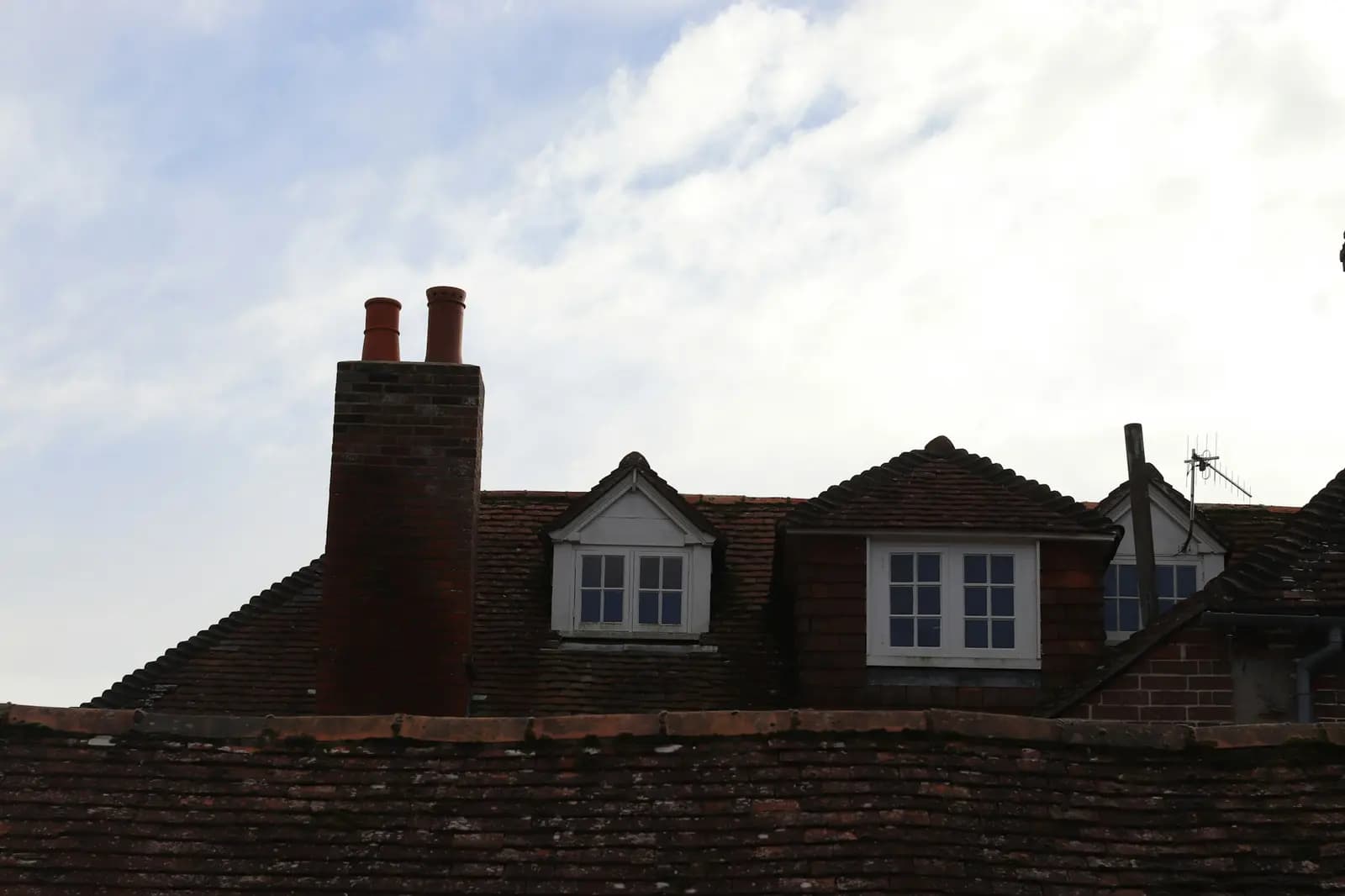 Clay-tiled roofs with a brick chimney and white dormer windows against a pale sky