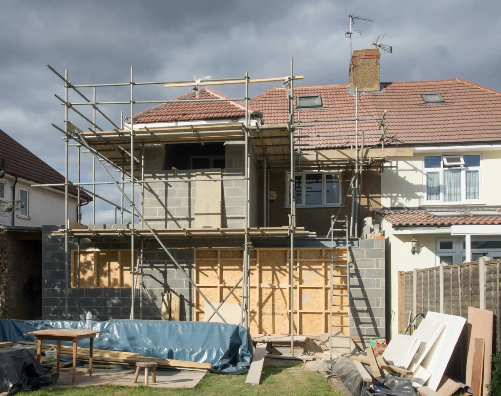 Rear of a semi-detached house with a two-storey extension under construction, showing exposed blockwork, timber boarding and scaffolding around the new walls