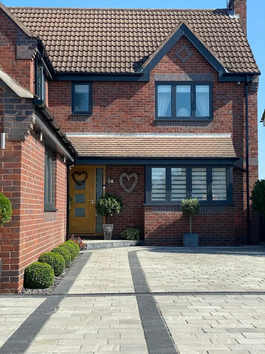 A red-brick detached house with a tiled roof and a yellow front door, with a block-paved driveway in two shades of grey edged by a dark linear drain