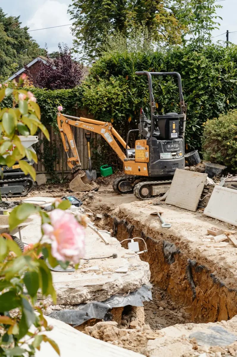 A mini excavator beside a deep trench and broken-out concrete slab in a back garden, with a timber fence, hedge and a brick house behind