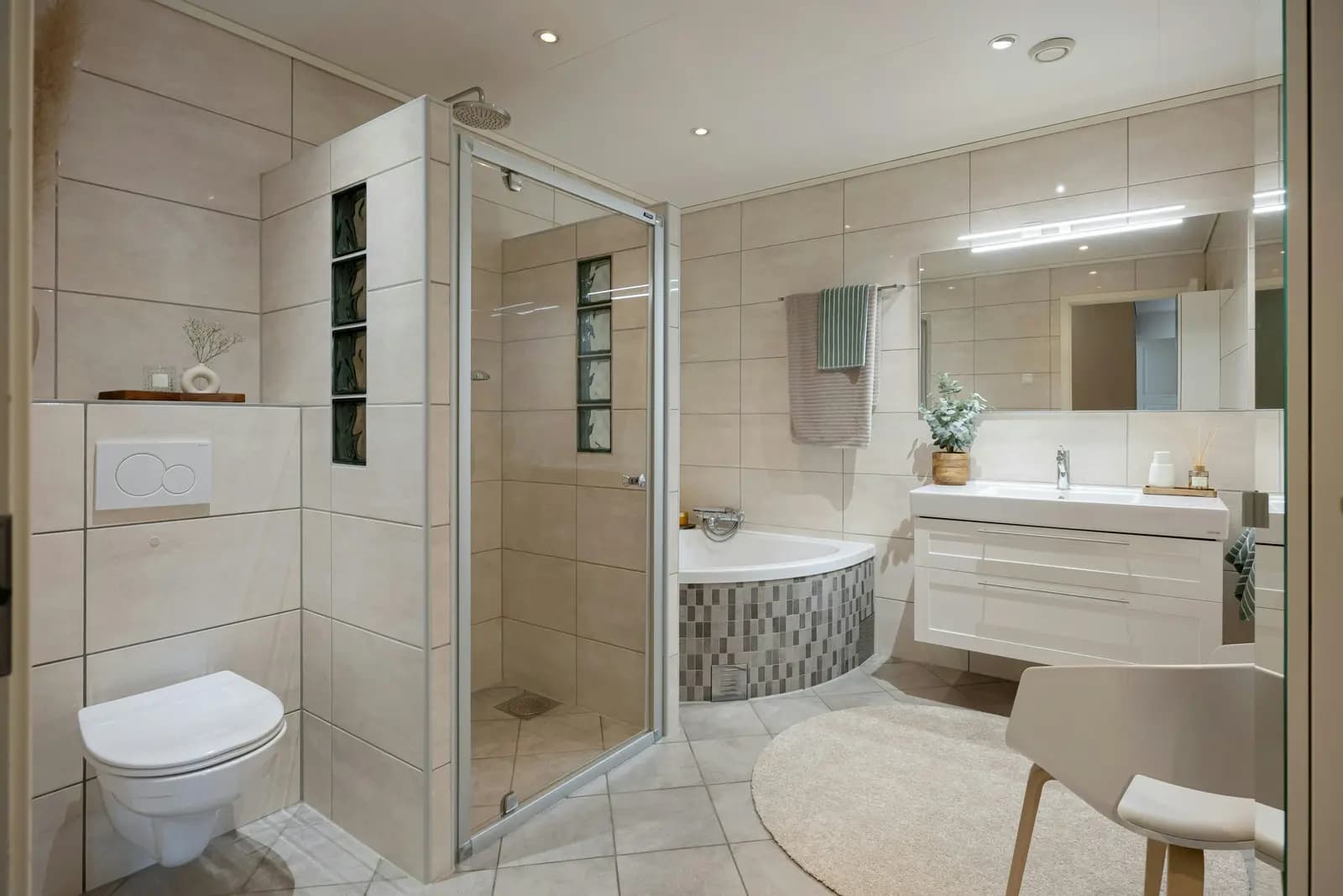 A fully tiled bathroom with a glass shower enclosure, corner bath, wall-hung toilet and a wall-hung vanity unit with a mirror and lights