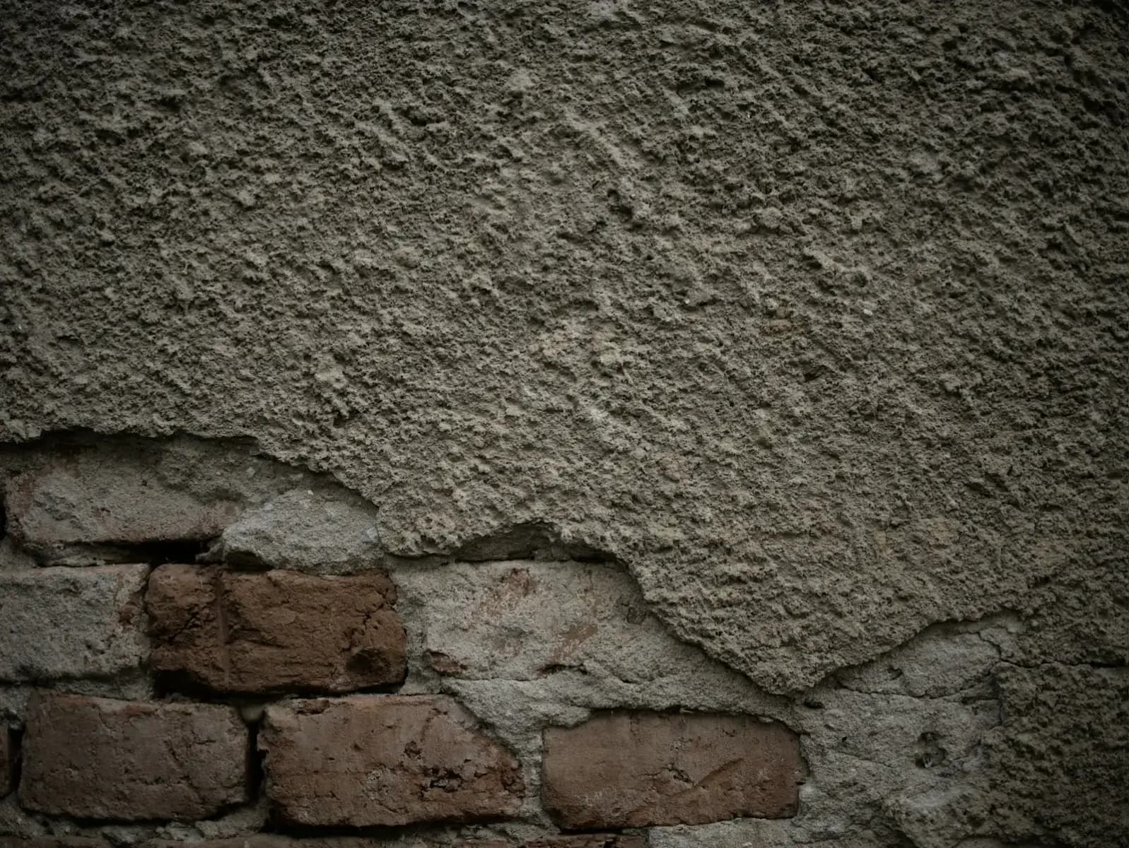 A rough, cement-coloured coating breaking away from a wall to show old brickwork underneath