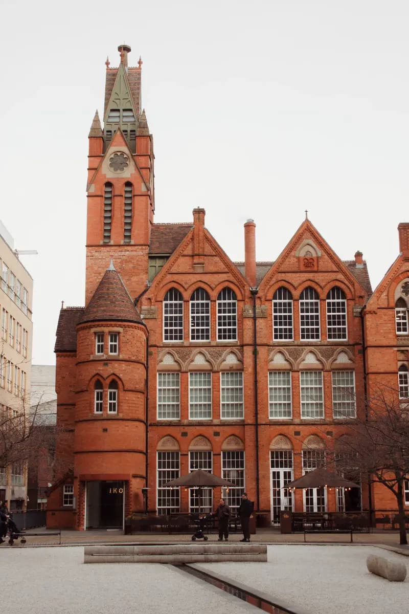 A Victorian Gothic red-brick building in Birmingham city centre with a tall pointed tower, a round turret, arched windows and a sign reading IKO above the entrance