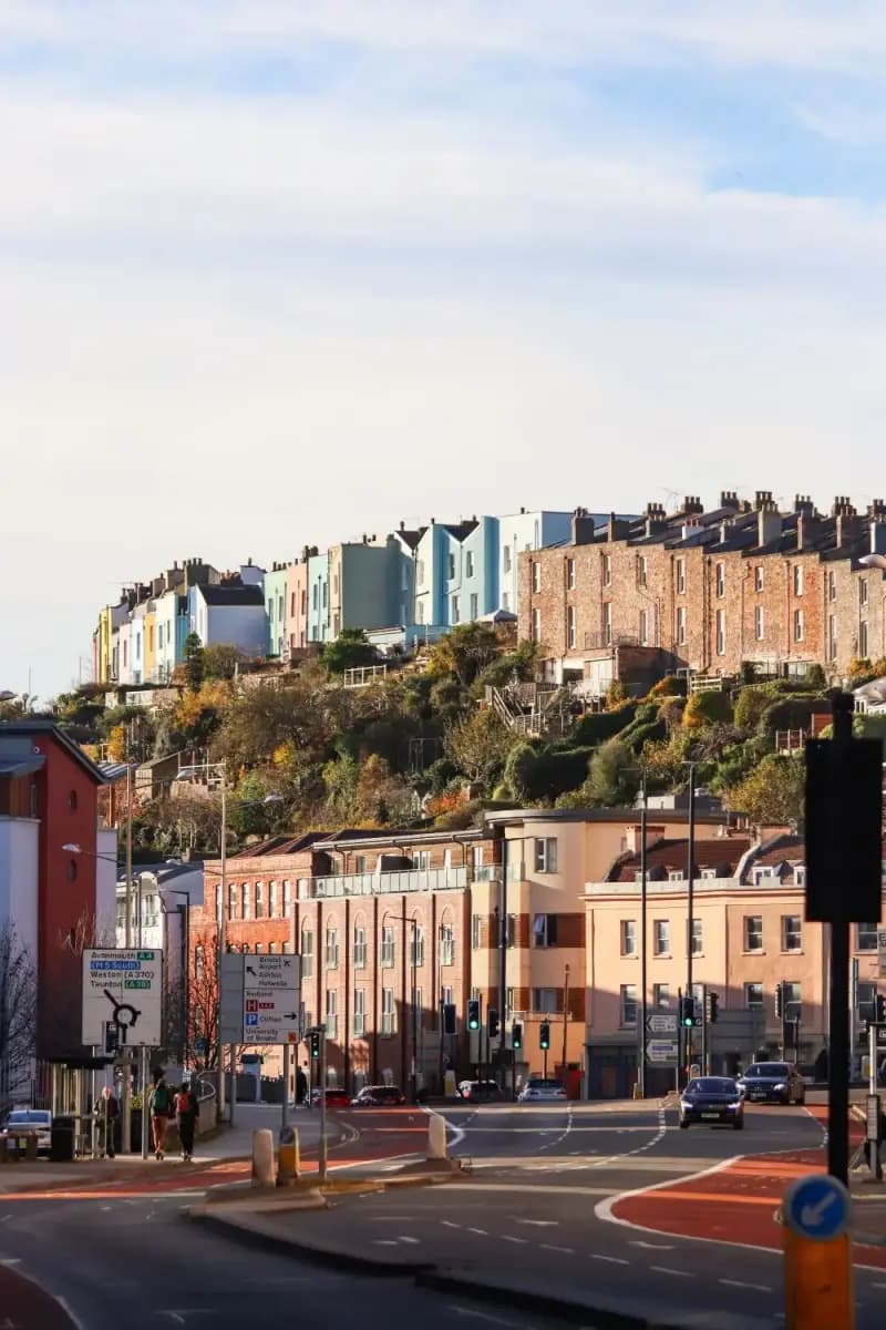 Painted Victorian terraces stacked on a Bristol hillside above a busy road, with a brick-built terrace to the right and UK road signs in the foreground