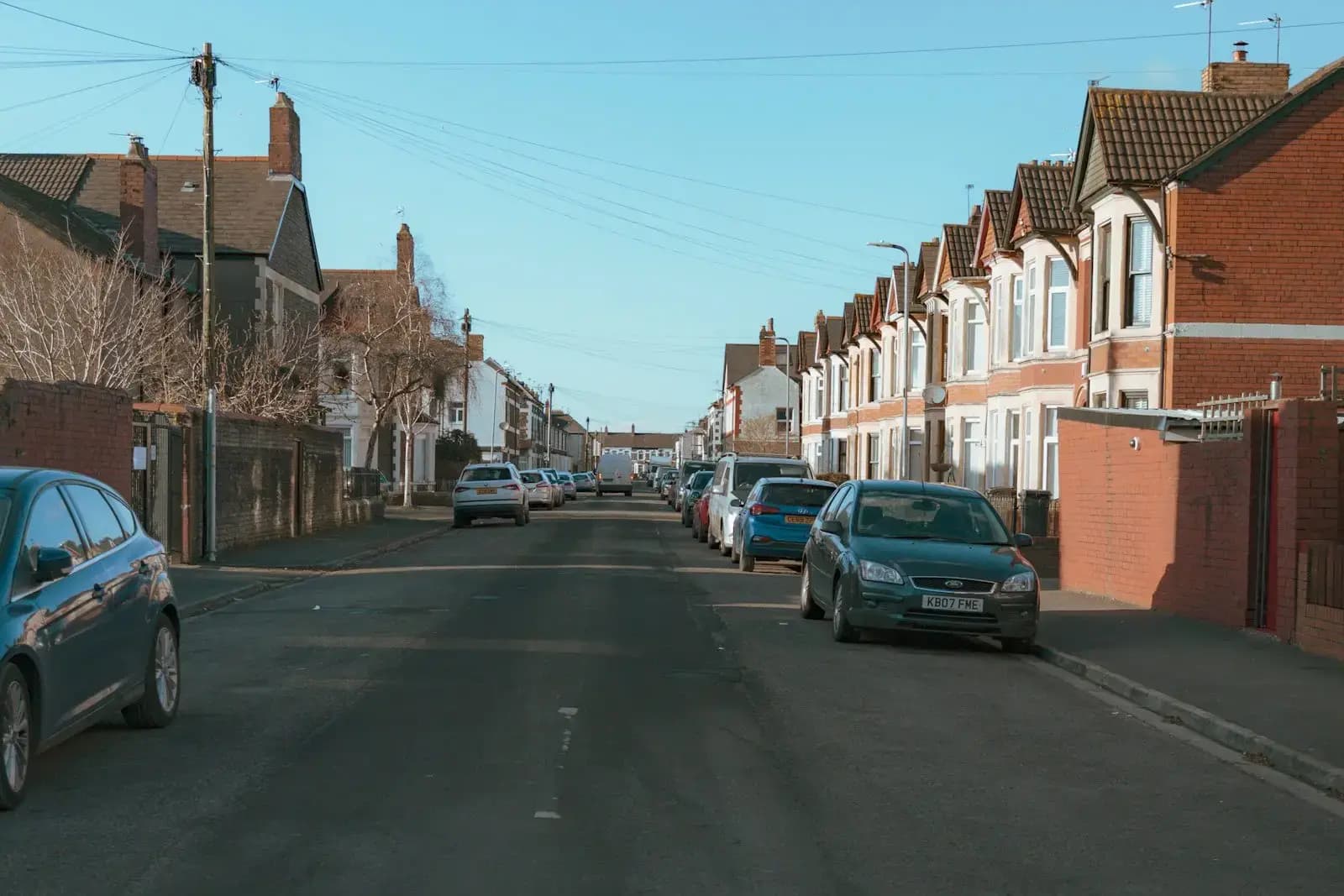 A Cardiff street of red-brick and rendered terraced houses with bay windows and tiled gables, cars parked along the kerb (cropped from the original photo)