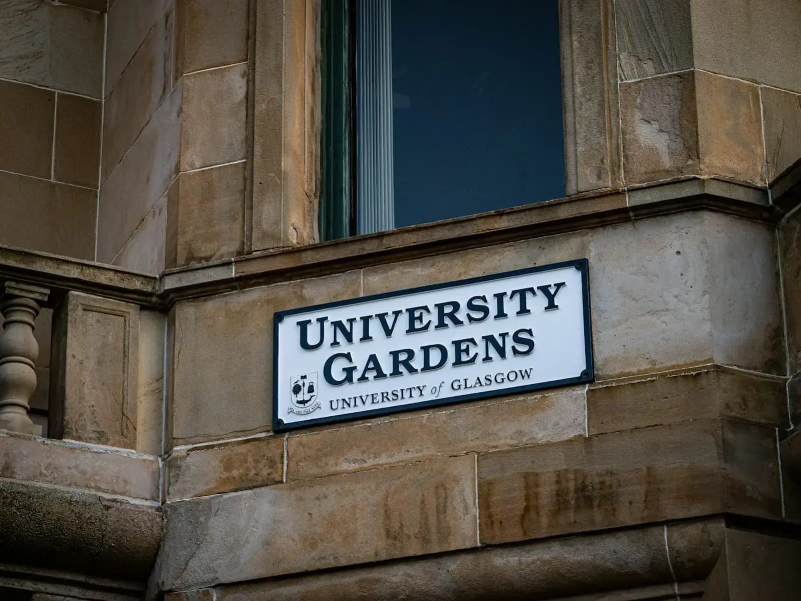 Close view of blond sandstone ashlar blocks and a bay window on a Glasgow tenement, with a University Gardens street sign