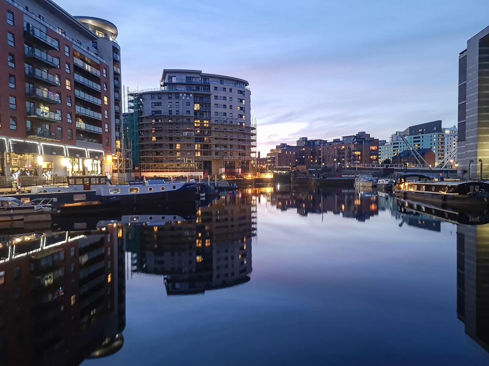 Leeds Dock at dusk with narrowboats moored along the quay and apartment blocks, one wrapped in scaffolding, reflected in the still water