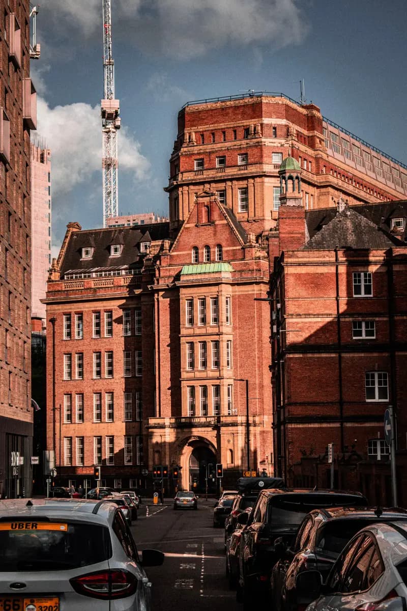 A street in Manchester city centre with traffic in the foreground and a large Edwardian red-brick building with an arched entrance, green copper details and a crane behind it