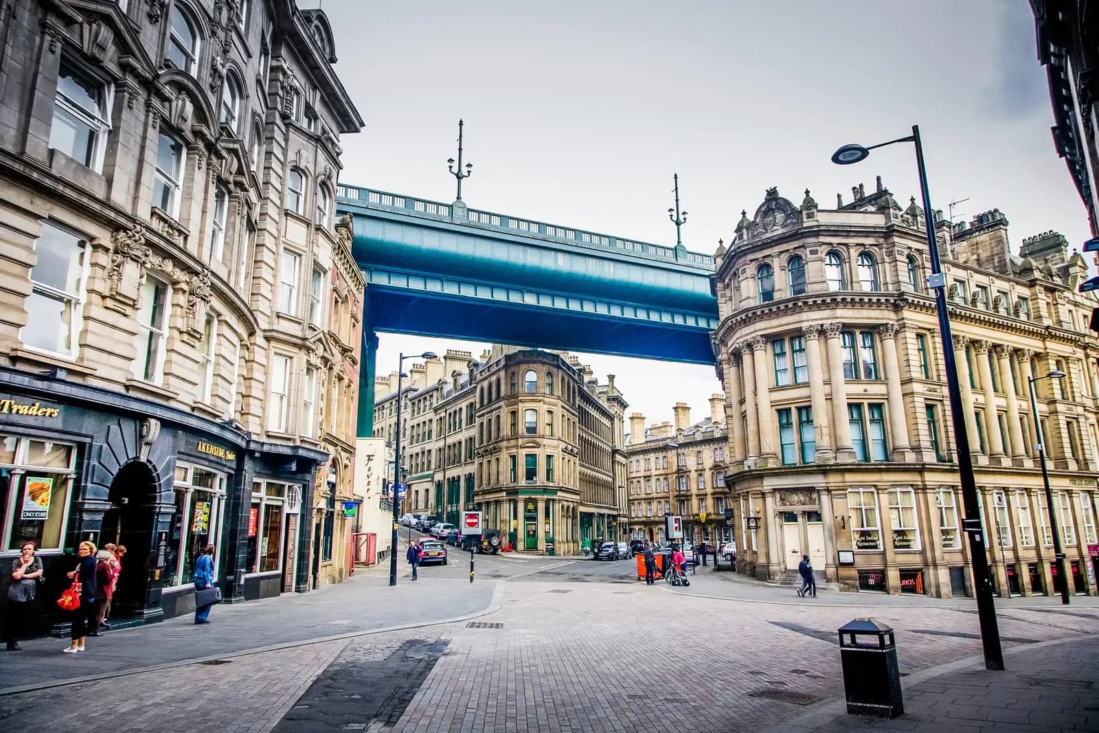 A street in central Newcastle lined with ornate stone Victorian buildings, with the High Level Bridge crossing overhead